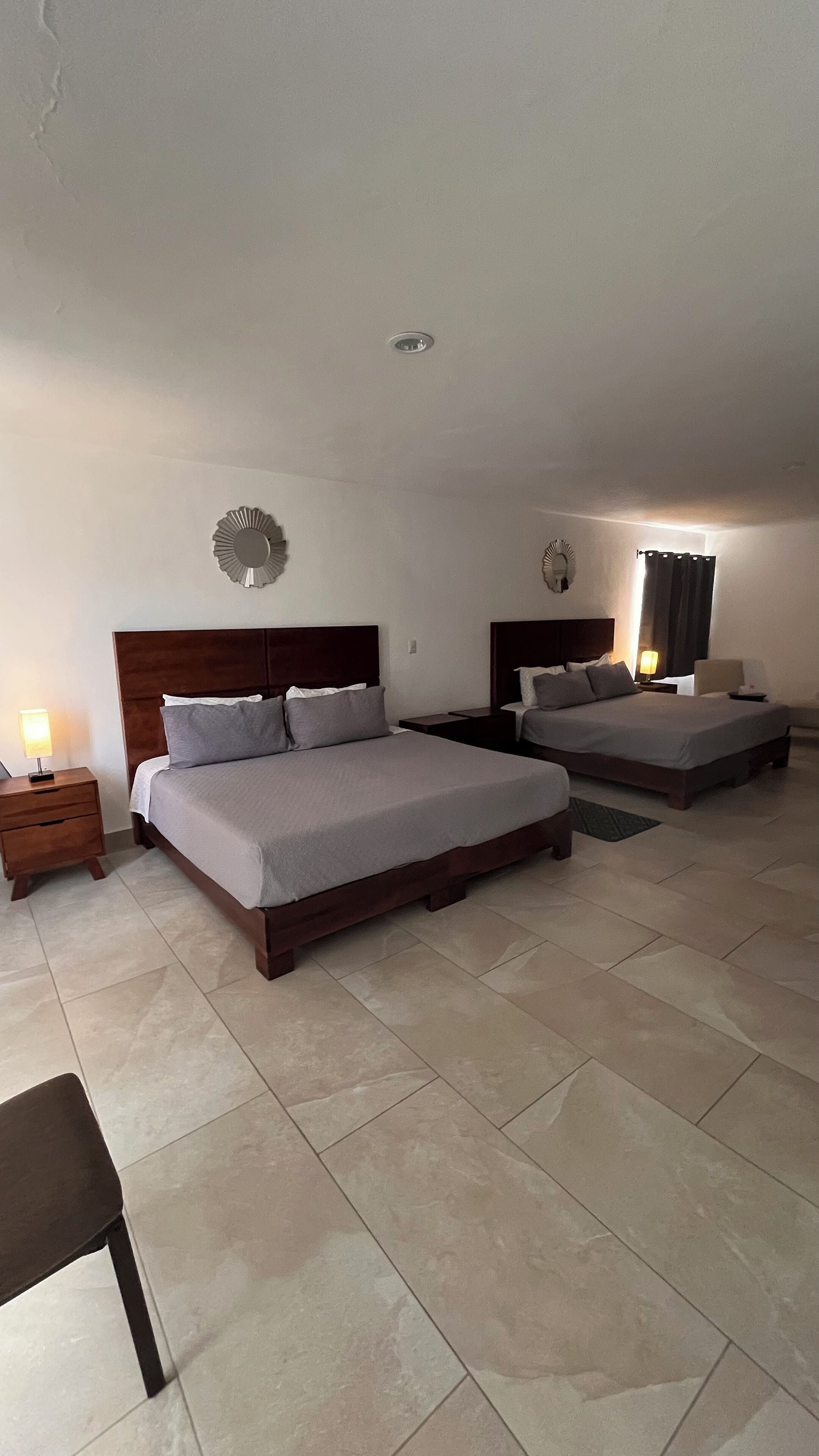 Quarto de hotel com duas camas, uma mesa de cabeceira com abajur e uma cadeira.