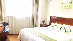 A neatly made bed with a green throw, a wooden nightstand with a vase of flowers, and a window with sheer curtains.
