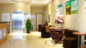 A hotel lobby with a reception desk, a chandelier, a plant, a round table with chairs, and informational posters on the walls.