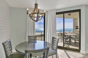A dining area with a round glass table, four chairs, and a chandelier. It features a view of the beach through large sliding glass doors.
