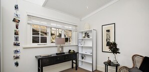 Une pièce comprenant une console noire, des étagères blanches, une œuvre d’art encadrée et un fauteuil en osier.