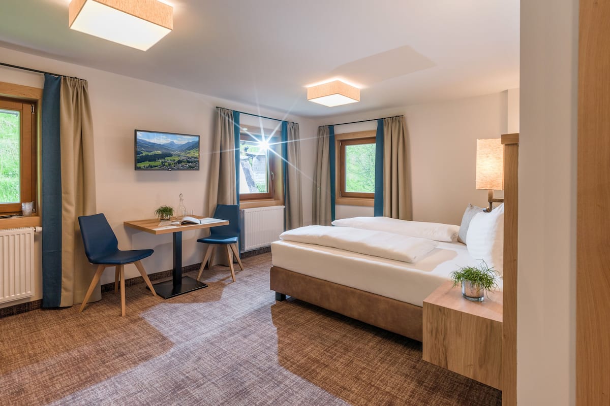 Una habitación de hotel con cama, escritorio con silla, ventana con cortinas, radiador y lámpara.