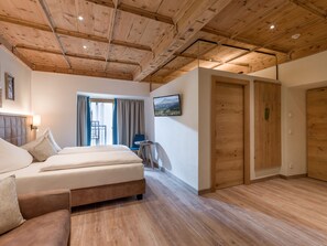 A hotel room with a wooden ceiling, a bed, a sofa, a small table, a chair, a window with curtains, and a door. - Alpen Glück Hotel Unterm Rain garni (Kirchberg in Tirol)