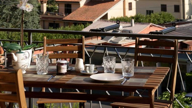 Une table à manger dressée avec un vase de fleurs, une théière et des verres, donnant sur un quartier résidentiel avec des maisons et une vue lointaine.