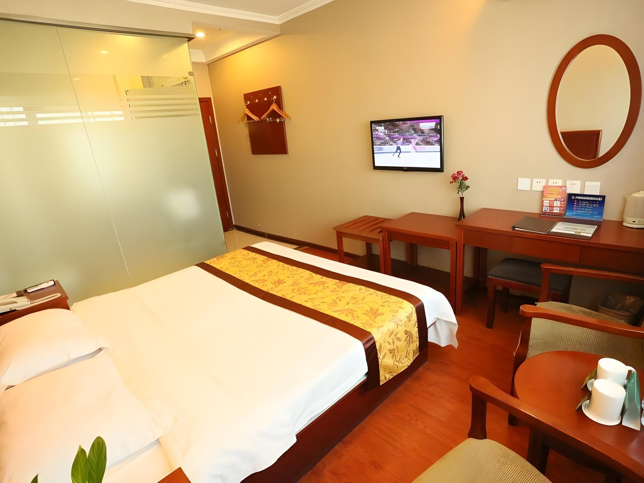 A hotel room with a bed, a desk, a chair, a small table with cups, a mirror, and a television on the wall.