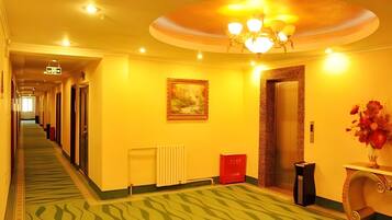 A hotel hallway with a green patterned carpet, a painting on the wall, a radiator, a red trash bin, an elevator, and a vase with flowers on a table.