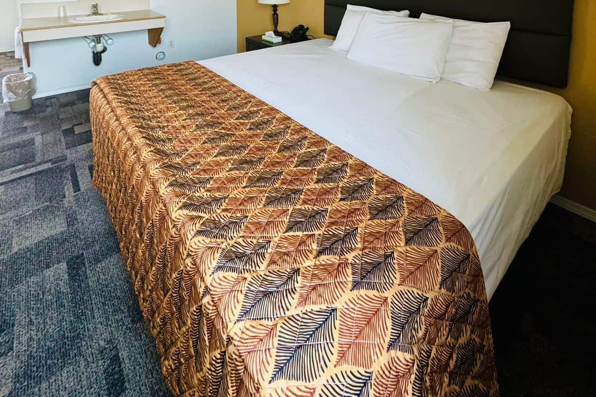 A hotel room with a bed, a bedside table with a lamp, a mirror, and a bathroom with a sink and mirror.