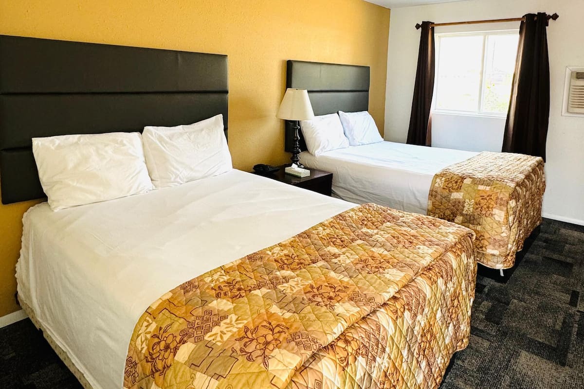 A hotel room with two beds, a headboard, a nightstand, a lamp, and a window with curtains.