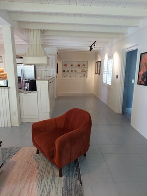 Un vaste espace de vie agrémenté d’un fauteuil rouge, une cuisine dotée de meubles blancs et une étagère murale ornée d’objets décoratifs.