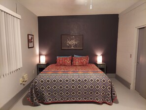 Une chambre à coucher avec un grand lit, deux tables de chevet avec des lampes, un tableau encadré au mur et une porte sur le côté droit.