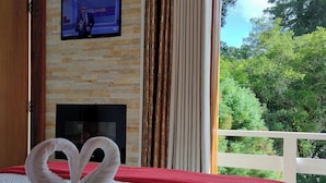 A hotel room with a stone wall, a flat-screen TV, a fireplace, and a balcony with a view of trees. - Casa do Papai Pousada (Campos do Jordão)