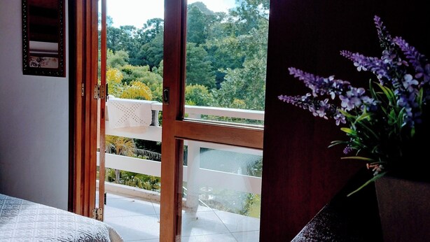 A room with a wooden ceiling, a sliding glass door leading to a balcony, and a vase with purple flowers on a table. - Casa do Papai Pousada (Campos do Jordão)