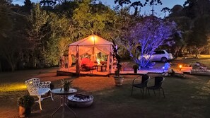 Un área al aire libre con carpas, asientos y un brasero, rodeada de árboles y un coche estacionado.