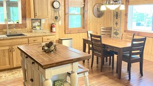 A cabin interior with a wooden dining table, chairs, a kitchen area with a sink, and a large window. - Experience the peaceful Wooded Retreat Cabin - 300 private acres -security gate  (Spencer)
