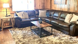 A cozy living room with a wooden ceiling, a leather sofa, a coffee table, and a large window. - Experience the peaceful Wooded Retreat Cabin - 300 private acres -security gate  (Spencer)