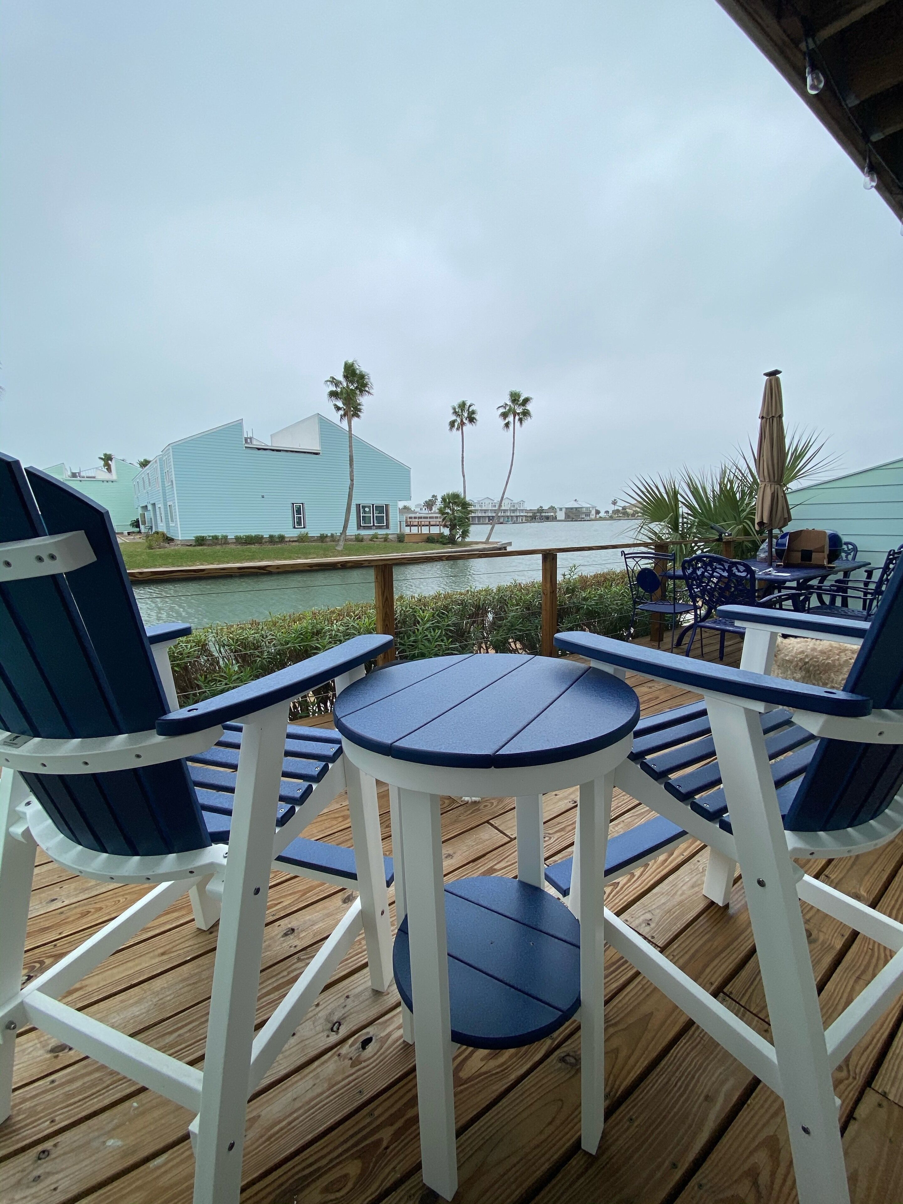 Eine Holzterrasse mit blauen und weißen Stühlen und einem kleinen Tisch, mit Blick auf ein Gewässer mit Gebäuden im Hintergrund.
