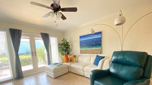 A living room with a ceiling fan, a teal armchair, a white sofa, and a painting of a beach scene. - Rockport(Little Palm@ Key Allegro)Waterfront Beautiful Condo Close to Everything (Rockport)