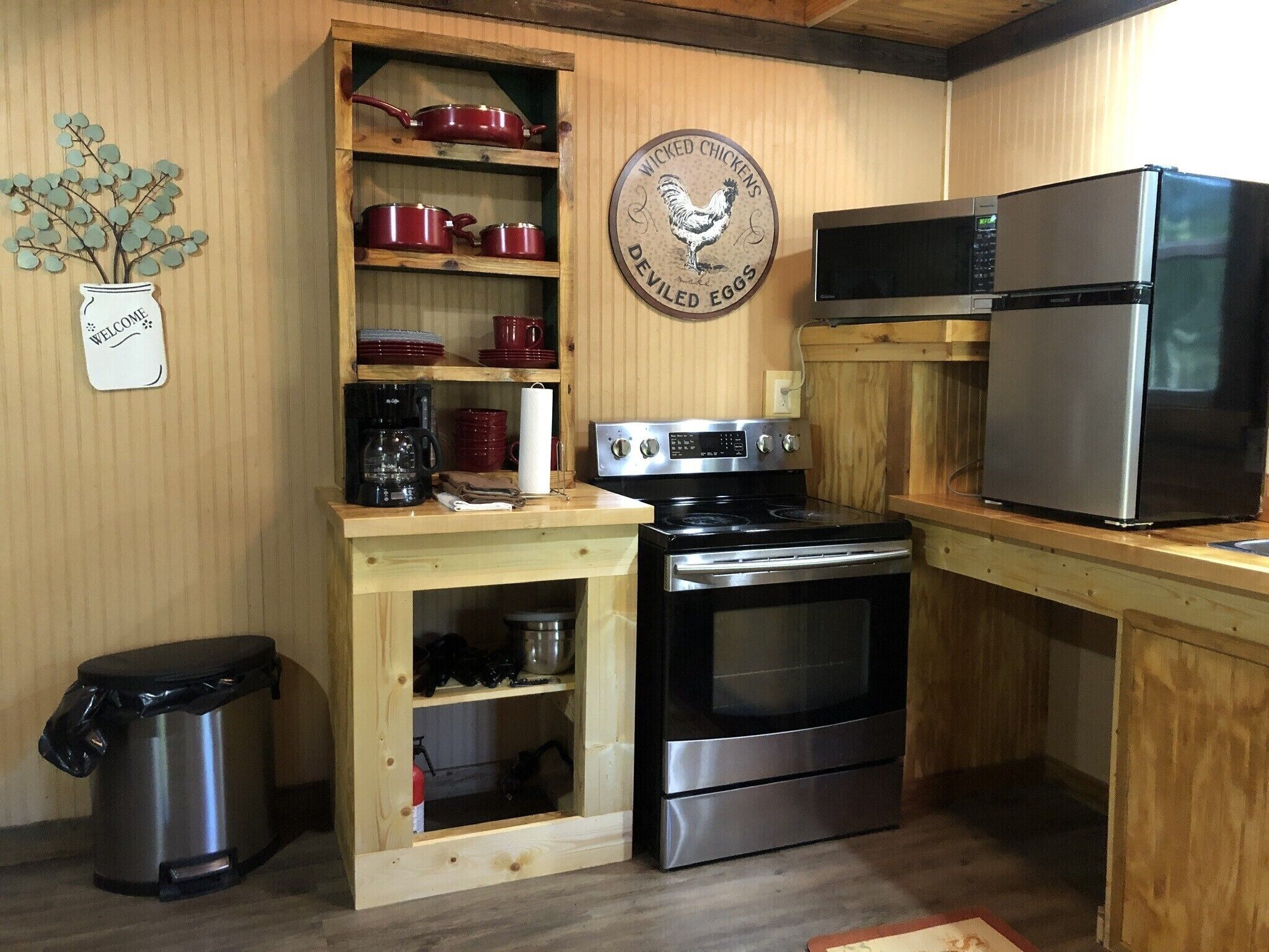 Una cocina compacta con estufa, horno y refrigerador. Se observa una estantería de madera con utensilios de cocina rojos y una cafetera. Hay un bote de basura a la izquierda.