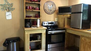 Una cocina compacta con estufa, horno y refrigerador. Se observa una estantería de madera con utensilios de cocina rojos y una cafetera. Hay un bote de basura a la izquierda.