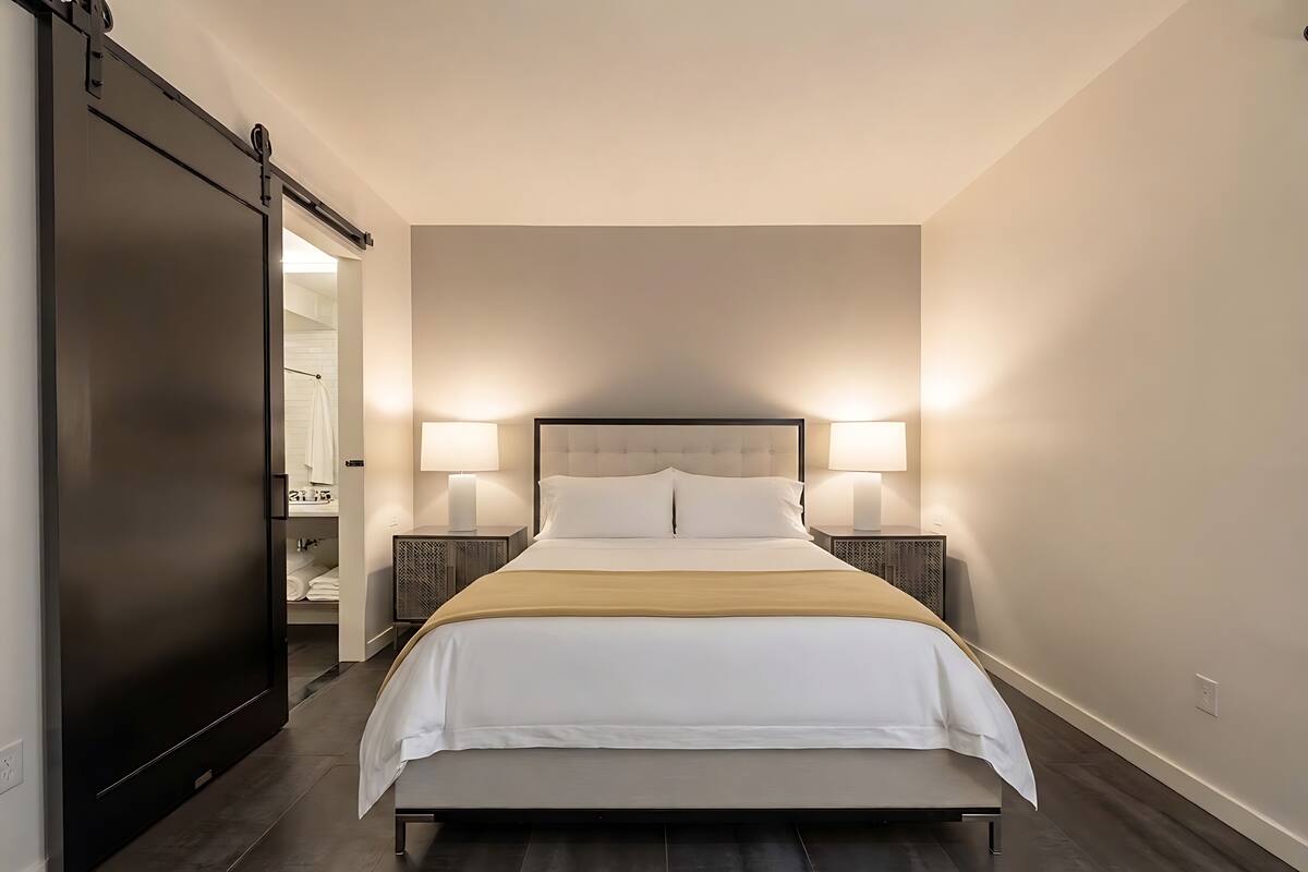 Habitació d'hotel amb llit, làmpades de sobretaula, una panxa i una porta corredissa que dona a un bany.