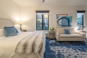 Una habitación de hotel con cama, sofá, un ventanal grande, un cuadro y una alfombra estampada.