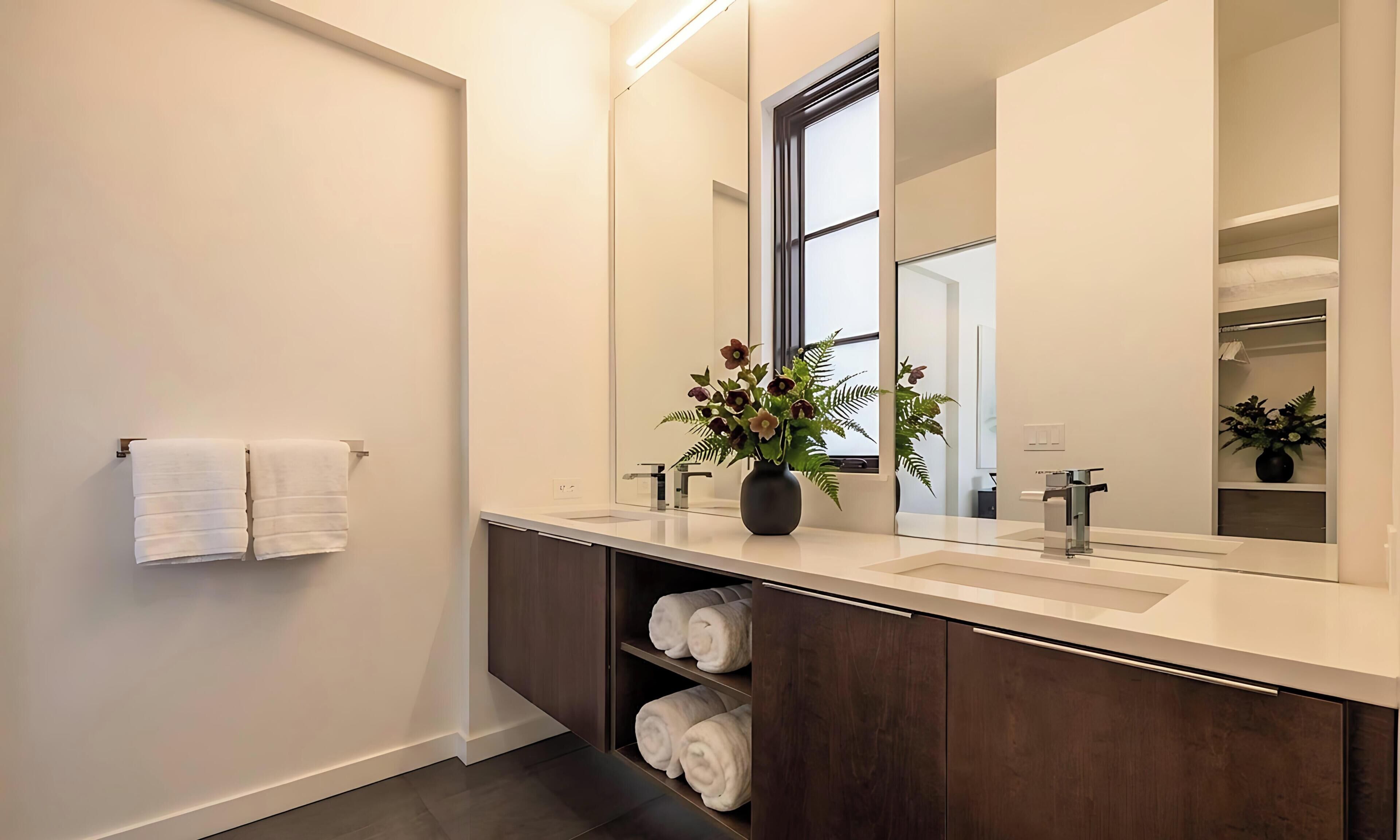 A modern bathroom with a large mirror, two sinks, a towel rack, and a plant.