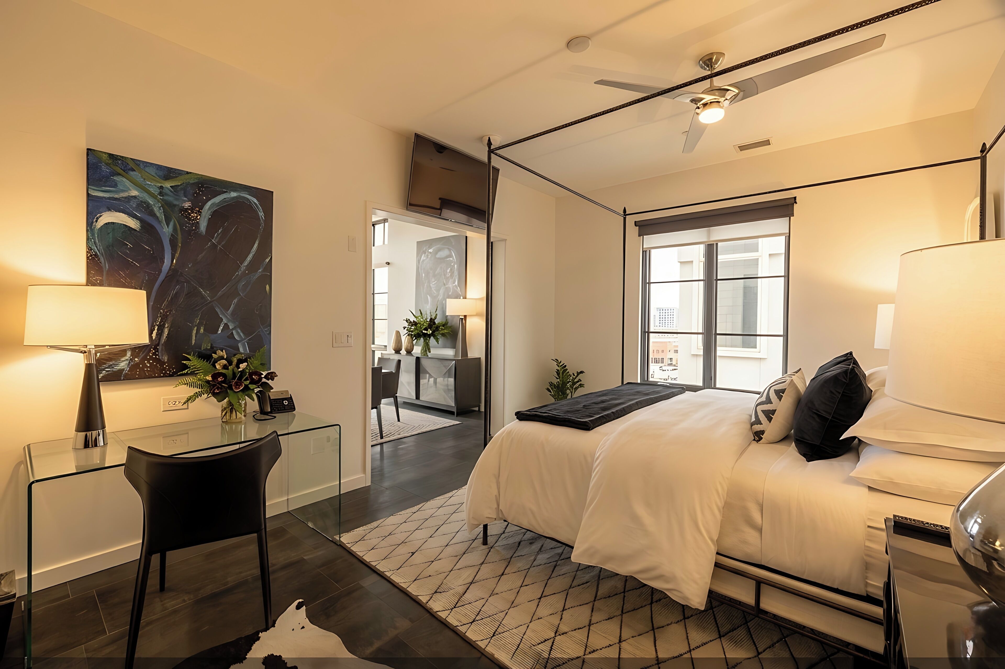 Una habitación de hotel moderna con un escritorio de vidrio, cama con ropa blanca, un ventanal grande y un ventilador de techo.