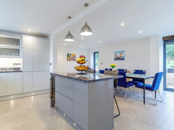 A modern kitchen with a central island, dining area with blue chairs, and artwork on the walls. - Skylark Barn-UK31354 (Swaffham)