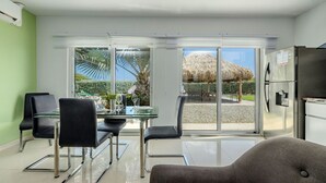Une cuisine moderne équipée d’un réfrigérateur, un coin repas avec une table et des chaises en verre, et une vue sur un patio doté d’une structure en chaume.