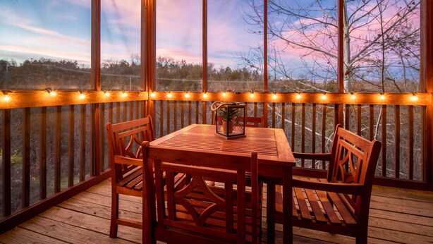 A wooden deck with a table and chairs, string lights, and a view of trees and sky.