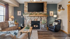 A cozy living room with a stone fireplace, a flat-screen TV mounted above it, a leather recliner, and a large window with curtains.