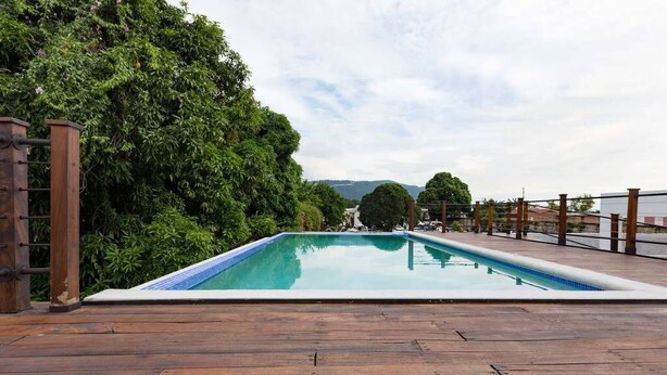 A rectangular swimming pool with a wooden deck in the foreground and dense greenery in the background. - The Brompton's @ Home (Kingston)