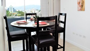 A dining area with a round table, black chairs, and a view of buildings through a large window. - The Brompton's @ Home (Kingston)