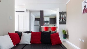 A modern living room with a black sofa, red and white cushions, a kitchen in the background, and a painting on the wall. - The Brompton's @ Home (Kingston)