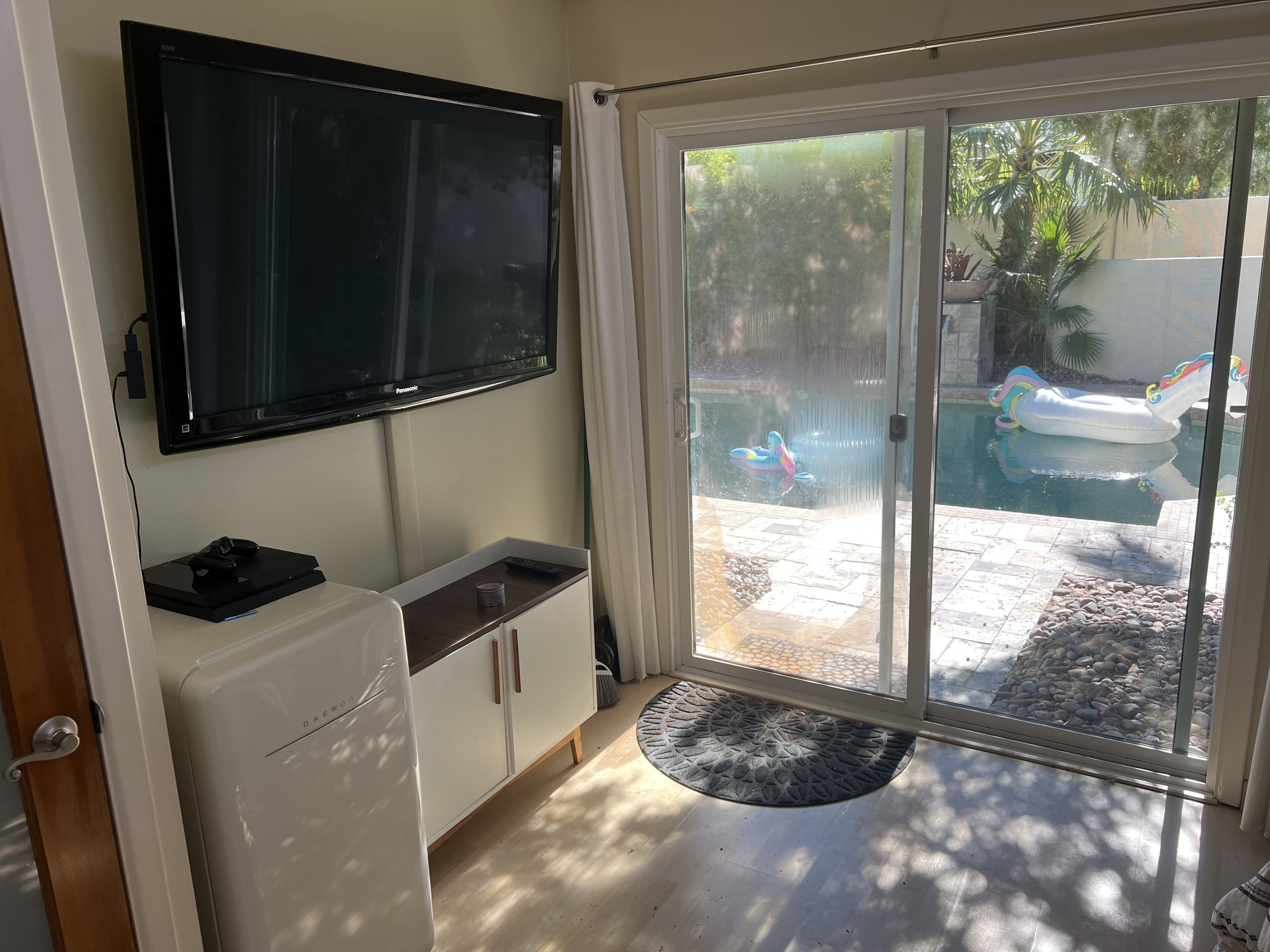 A room with a flat-screen TV, a small refrigerator, a cabinet, and a sliding glass door leading to a pool area with inflatable toys.
