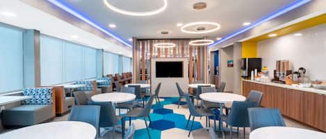 A modern dining area with round tables and blue chairs, a flat-screen TV, and a kitchen counter with appliances.