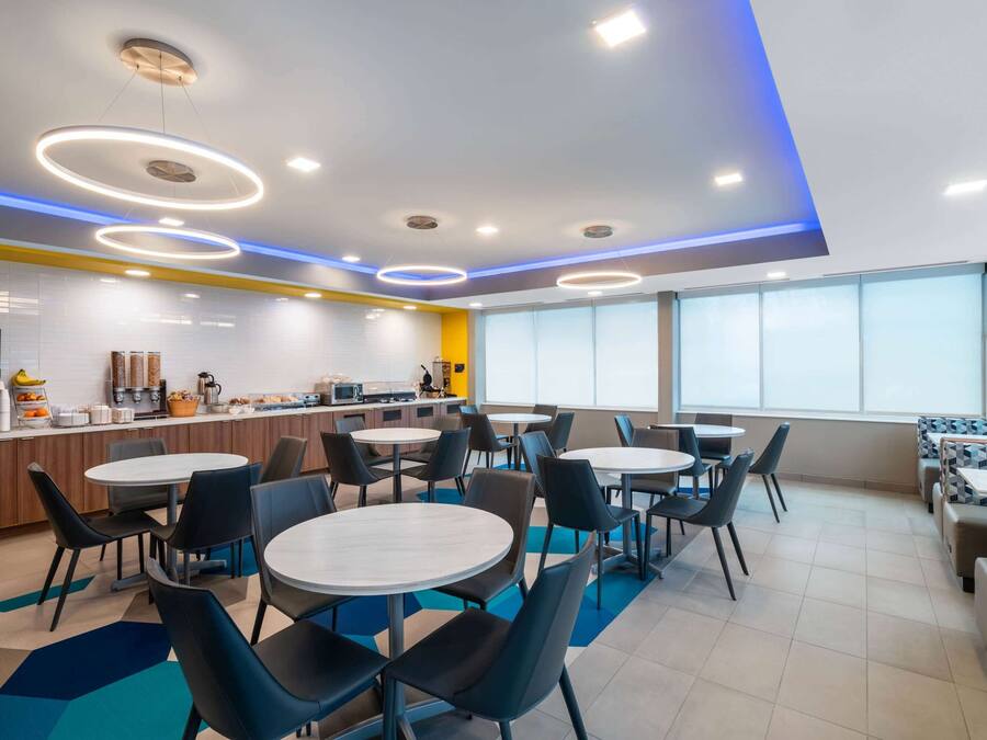 A modern dining area with round tables and chairs, a counter with a coffee machine, and a large window providing natural light.