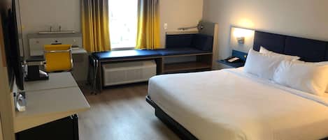 A hotel room with a bed, desk, chair, and a small seating area by the window.