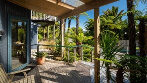 Eine Holzterrasse mit Glastür, die zu einem Raum führt, umgeben von tropischen Pflanzen und blauem Himmel.