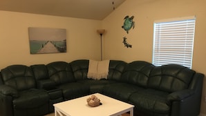 Ein Wohnzimmer mit einem dunkelgrünen Ledersofa, einem weißen Couchtisch, einem Gemälde mit einer Küstenansicht, einer Stehlampe und einem Fenster mit Jalousien.