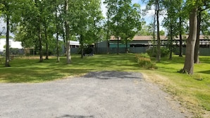 A gravel driveway leading to a grassy area with trees, a fenced yard, and a building with a metal roof. - The Keuka at Red Brick Manor (Seneca Falls)
