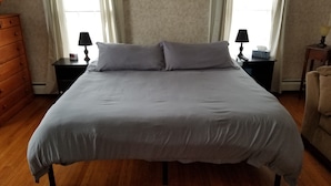A neatly made bed with a grey bedspread and two pillows, flanked by nightstands with lamps and a clock. A wooden dresser is to the left, and a chair is to the right. - The Keuka at Red Brick Manor (Seneca Falls)
