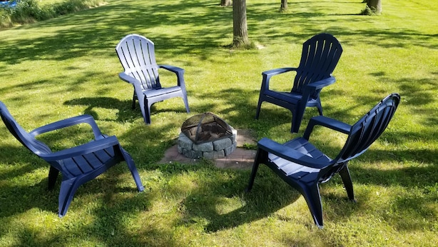 A circular arrangement of four Adirondack chairs around a stone fire pit on a grassy lawn. - The Keuka at Red Brick Manor (Seneca Falls)