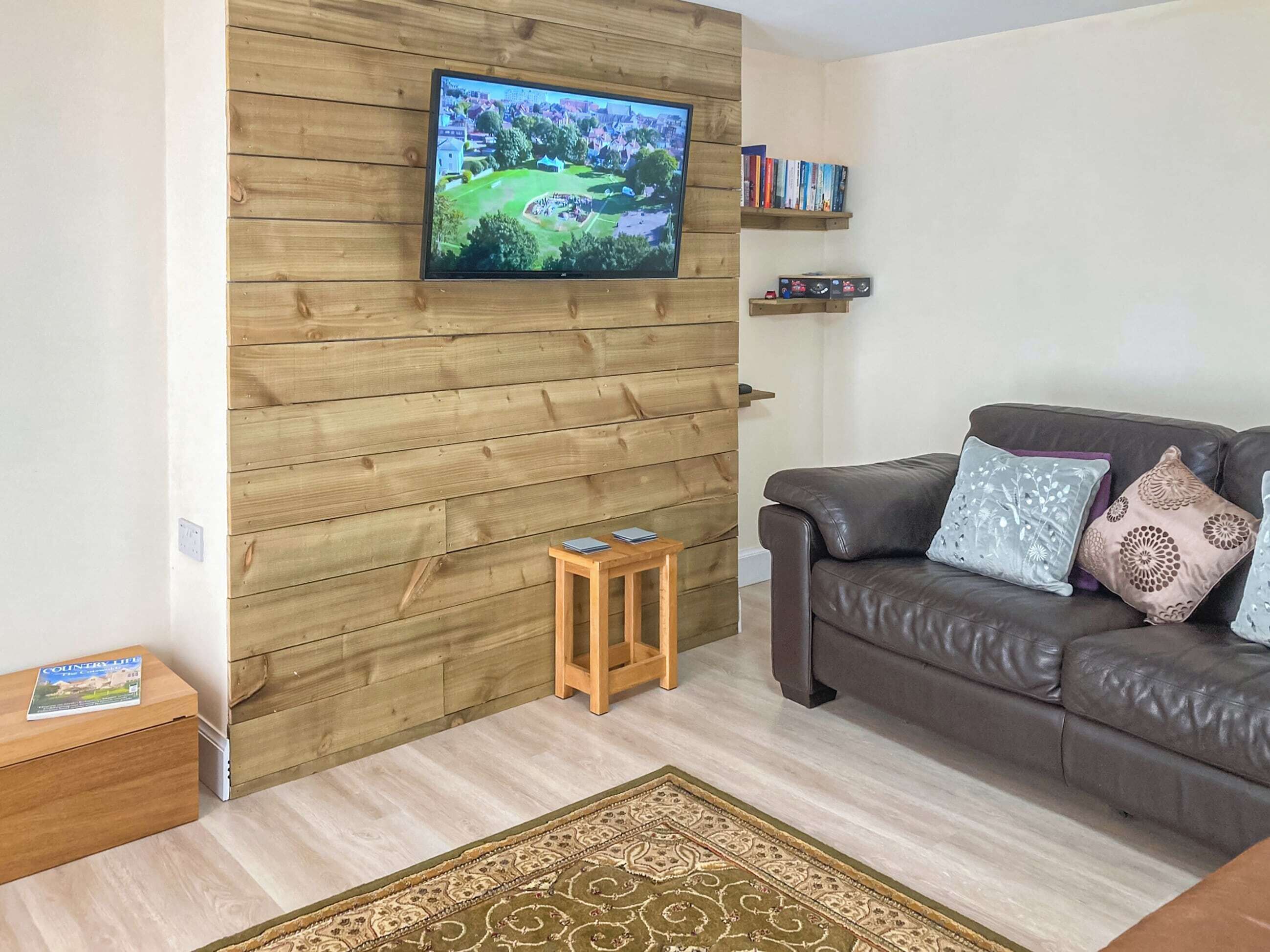 Un salon avec un mur en bois, un téléviseur fixé au mur, un canapé en cuir marron agrémenté de coussins à motifs, une petite table d’appoint en bois et un tapis décoratif.