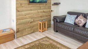 Uma sala de estar com parede de madeira, uma televisão montada nela, um sofá de couro marrom com almofadas estampadas, uma pequena mesa lateral de madeira e um tapete decorativo.