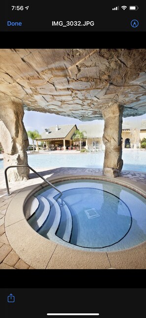 Un bain à remous sous une structure en pierre, avec une piscine et un bâtiment en arrière-plan.