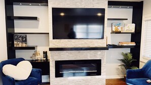 Una sala de estar moderna con chimenea, dos sillones azules y un televisor montado en una pared de ladrillo.