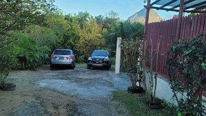 Un camino de grava conduce a una casa con dos coches aparcados, rodeada de abundante vegetación y una montaña al fondo.