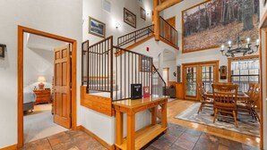 A spacious interior with a wooden staircase, a dining area with a large painting, and a view through the windows. - Walk-In 2BR Grand Mountain Cabin with Ozark Views | Sleeps 6 (Branson)