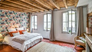 A bedroom with a wooden ceiling, a floral wallpaper, a bed with white bedding, and a window with curtains.
